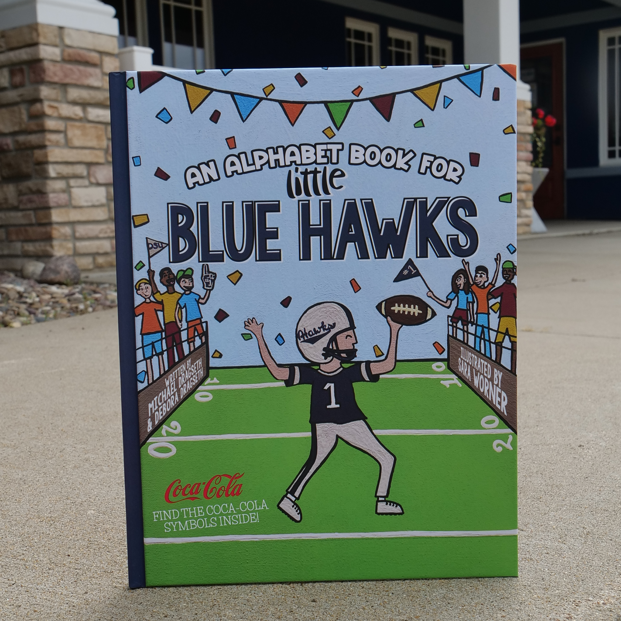 Children's book titled 'An Alphabet Book for Little Blue Hawks' on a sidewalk.