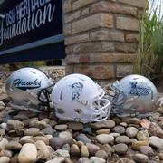 Three miniature football helmets on pebbles with a stone building and sign in the background