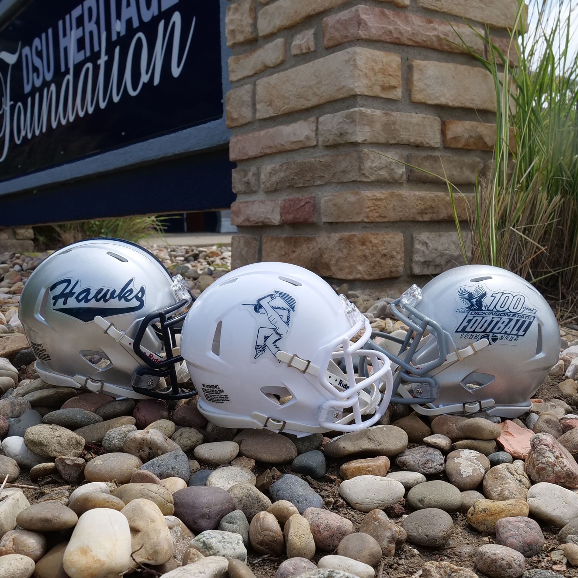 Three miniature football helmets on pebbles with a stone building and sign in the background