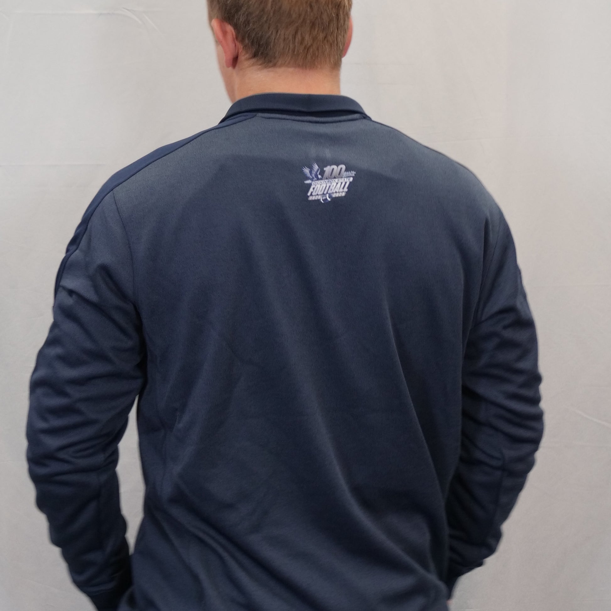 Person wearing a navy blue jacket with a logo on the back against a light gray wall